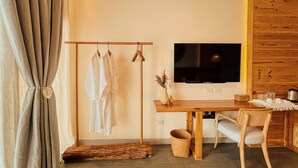 A room with a wooden TV stand, a dining table with chairs, a wooden floor, a clothes rack with white garments, and a large window with curtains. - Molly Resort Bohol (Panglao)