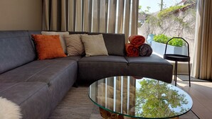 Ein modernes Wohnzimmer mit einem Ecksofa, einem Glastisch und Blick auf einen Garten durch große Fenster.