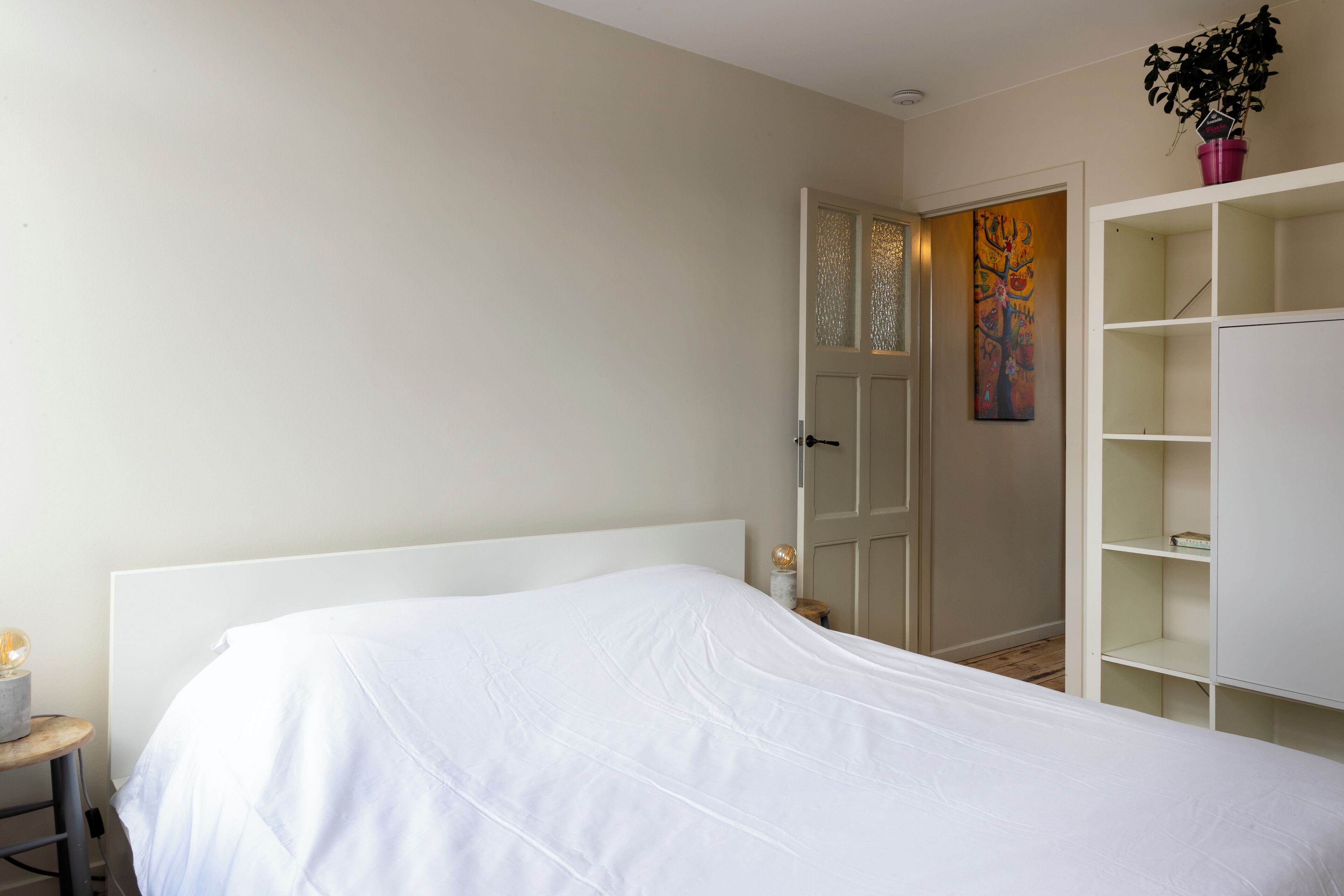A bedroom with a white bed, a nightstand with a lamp, a door with frosted glass panels, and a white shelving unit.