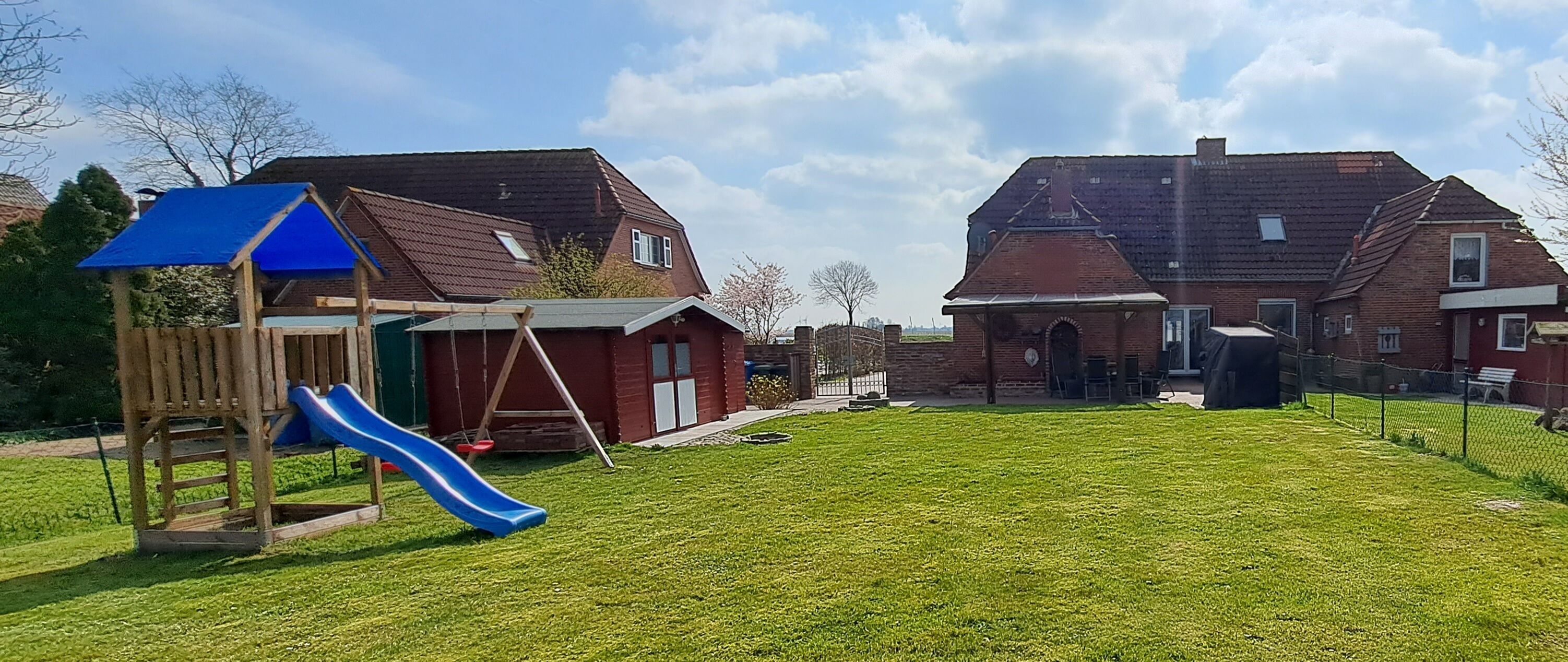 Un patio trasero con un parque infantil de madera, un tobogán azul y una casa de ladrillo con techo rojo.
