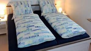 En dubbelsäng med en vit sängram och blått sänglinne, två sänglampor, en tavla som föreställer en strandscen och en taklampa.
