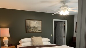 Een slaapkamer met een bed, nachtkastje, lamp, plafondventilator, deur en een ingelijst schilderij aan de muur.