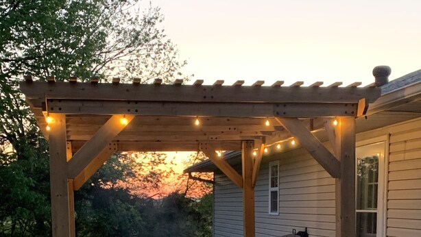 Een terras buiten met twee Adirondack-stoelen en een vuurkuil, onder een houten pergola met lichtjes.