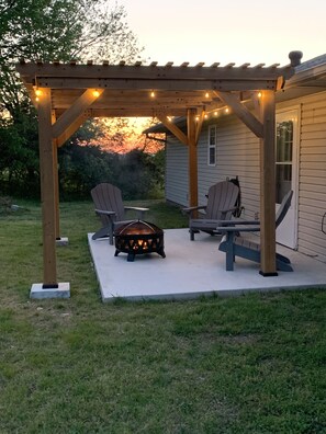 Une terrasse extérieure avec deux fauteuils Adirondack et un foyer, située sous une pergola en bois illuminée de guirlandes lumineuses.