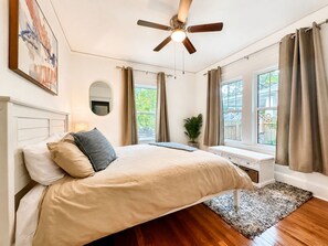 Un dormitorio con cama, ventilador de techo, ventana con cortinas, mesita de noche y una planta.
