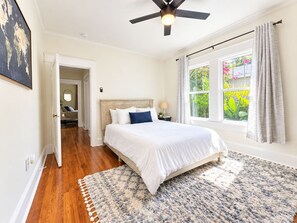 Quarto com cama, ventilador de teto, janela com cortinas e vista para o verde lá fora.