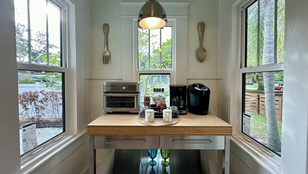 Una cocina con encimera de madera, cafetera, tostadora y ventana con vista a árboles y una valla.