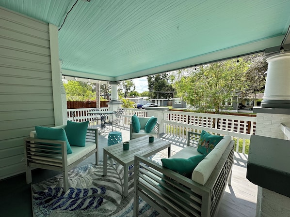 A covered outdoor patio with white furniture, a patterned rug, and a view of the neighborhood.