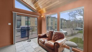 A room with a brown leather sofa, a glass door leading to an outdoor patio with a barbecue grill, and a view of a parking lot and buildings outside.