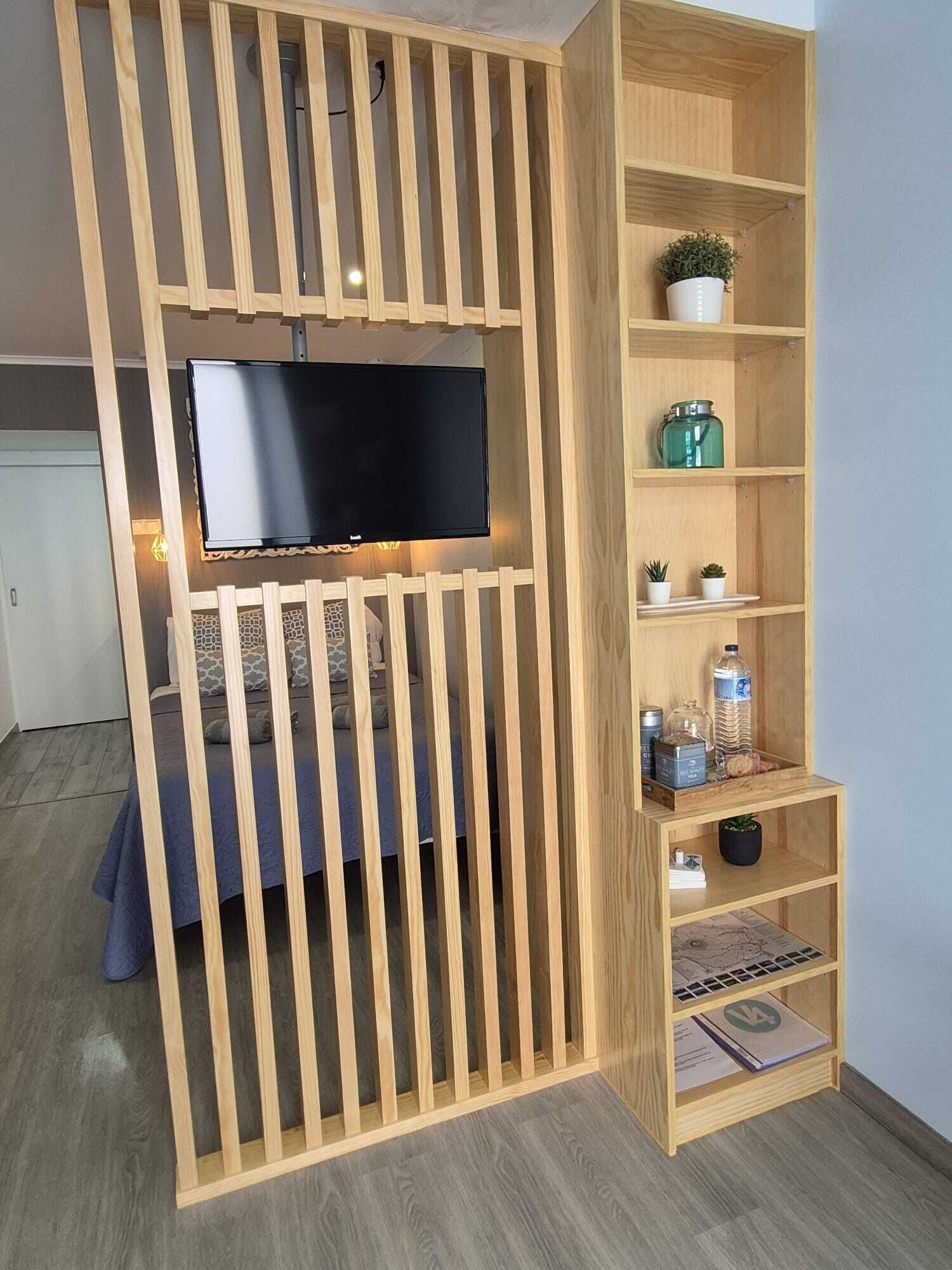 Habitació amb televisió penjada a una separació de fusta, un llit i una prestatgeria amb diversos objectes.
