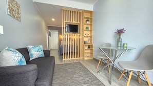 Una sala de estar moderna con un sofá gris, una mesa de comedor de vidrio con patas metálicas y una separador de madera con estantes.
