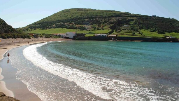 Una playa con una costa curvada, un edificio pequeño y una colina al fondo.