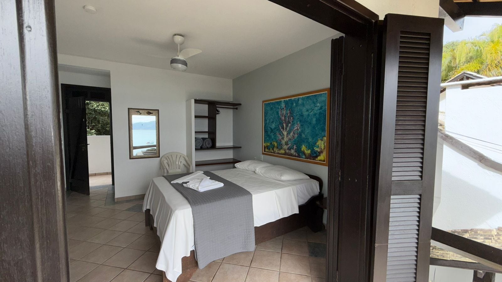 Une chambre à coucher avec un lit, une chaise, un ventilateur de plafond, un tableau et une vue sur l’extérieur.