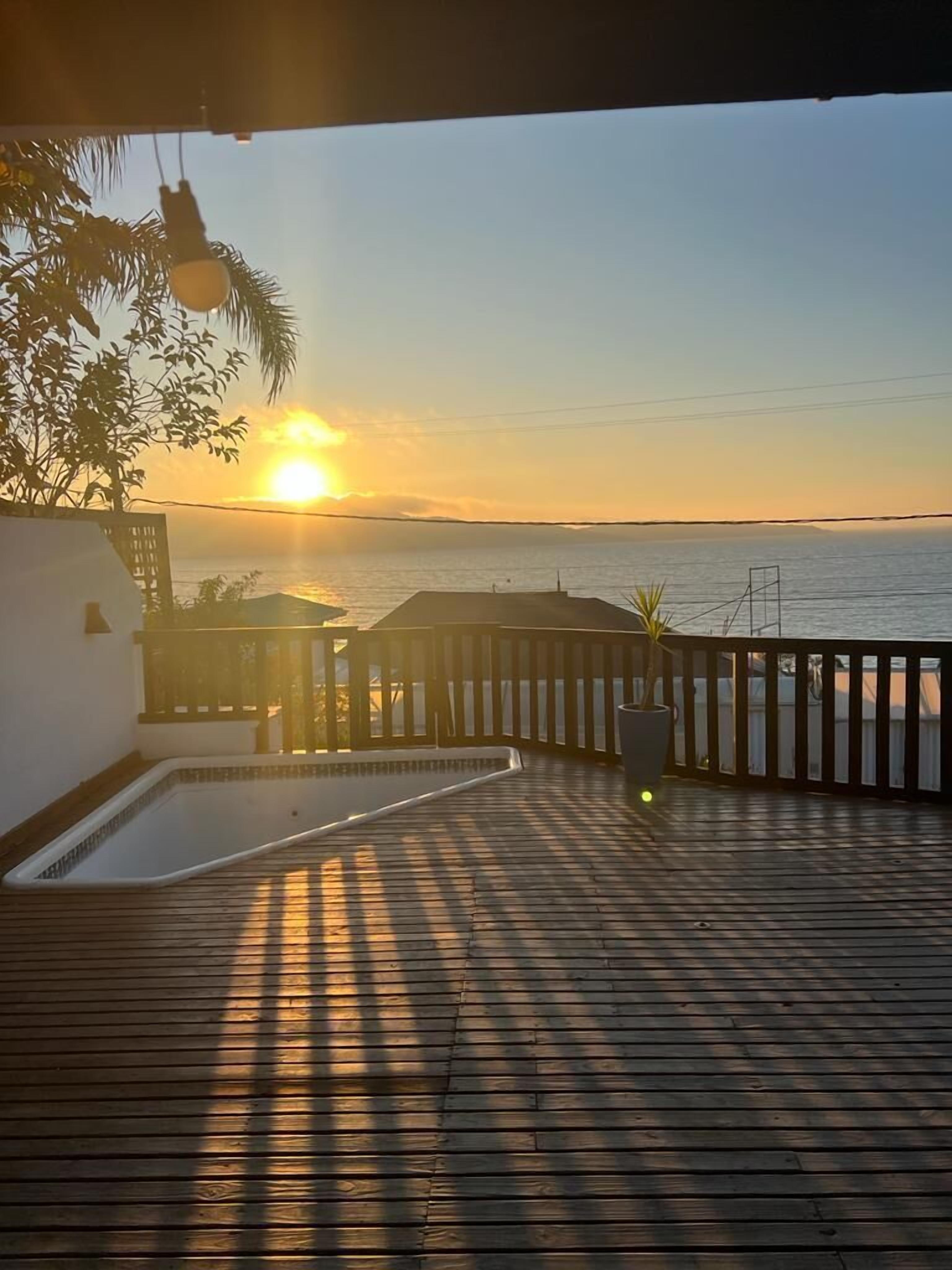 A wooden deck with a pool, overlooking a sunset over the ocean.