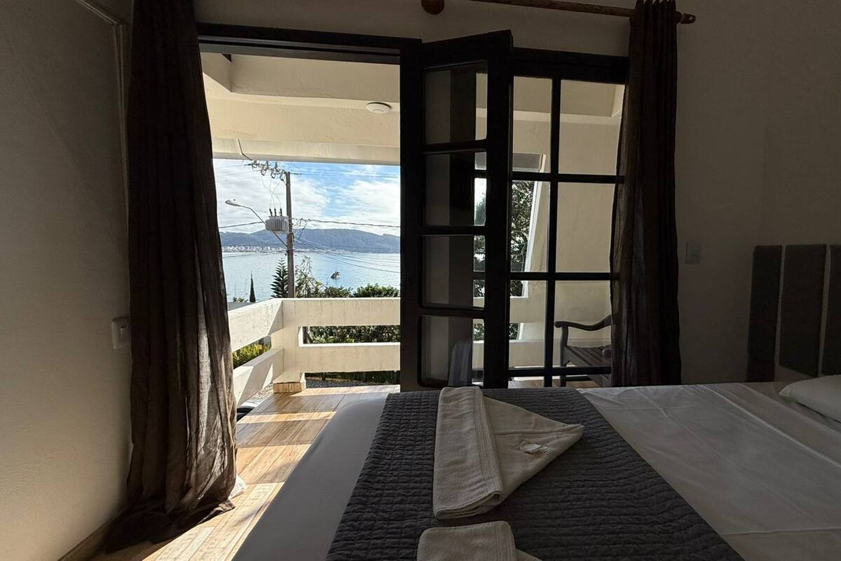 A hotel room with a bed, a view of the sea, and a balcony with a chair.