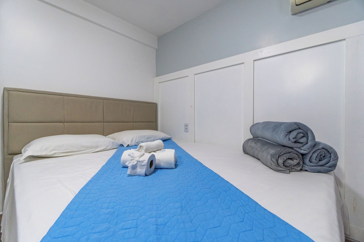 A neatly made bed with a blue quilt, white pillows, and rolled towels on the side.
