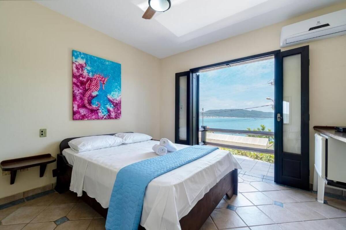 A bedroom with a bed, a painting, a bench, and a view of the sea through an open door.
