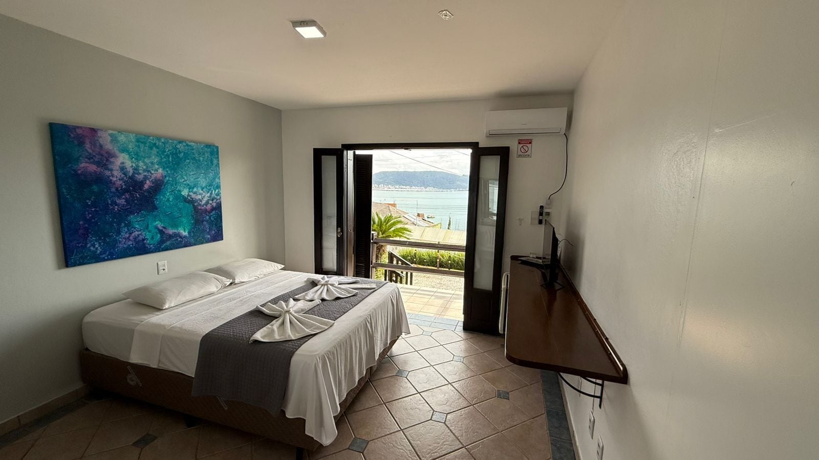 A hotel room with a bed, a painting on the wall, an open door leading to a balcony with a view of the sea, and an air conditioning unit.