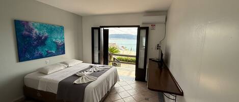 A hotel room with a bed, a painting on the wall, an open door leading to a balcony with a view of the sea, and an air conditioning unit.