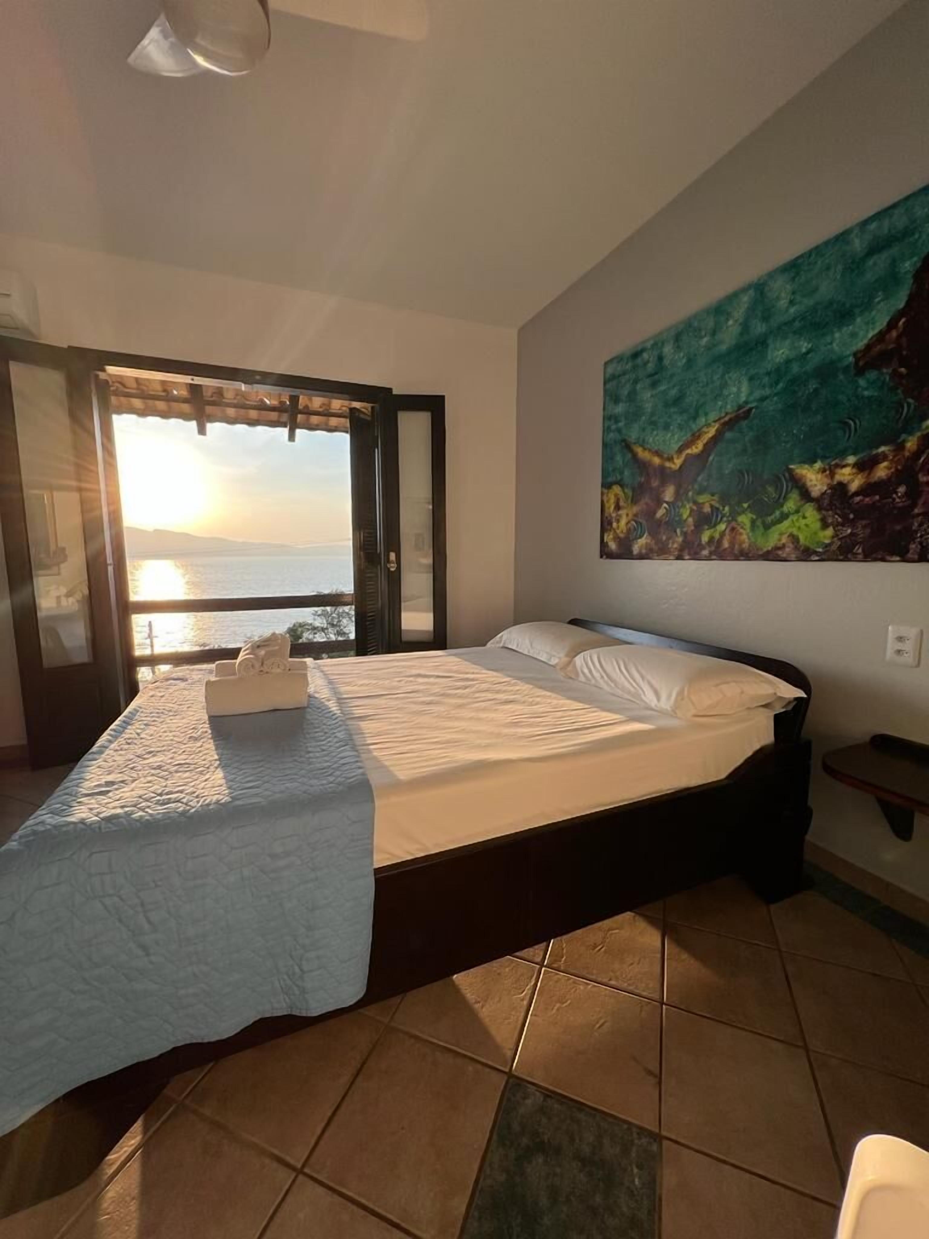 A bedroom with a large bed, a view of the sea through a sliding door, and a painting on the wall.