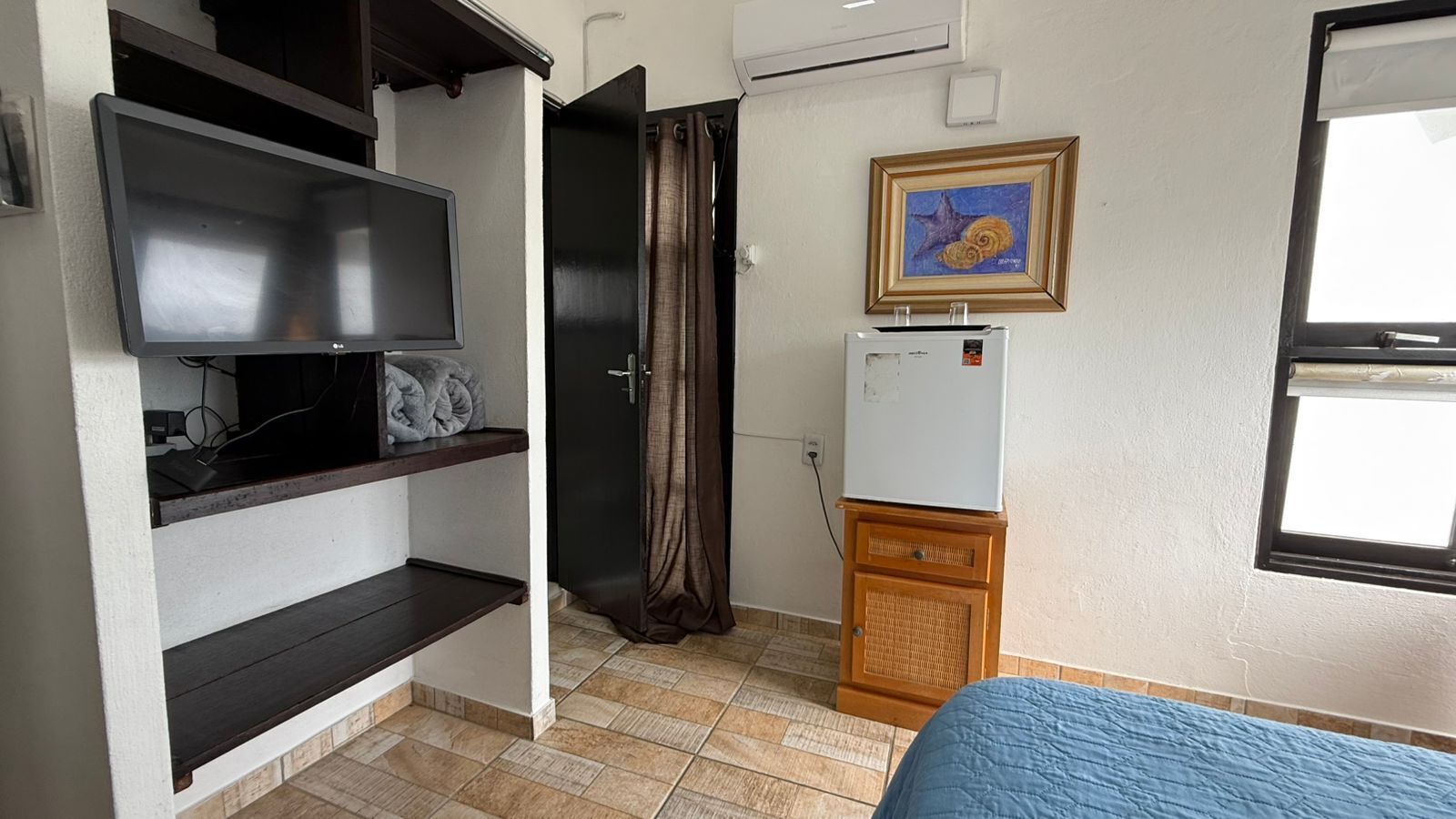 A compact room with a bed, a TV on a shelf, a small refrigerator, and a window with a curtain.