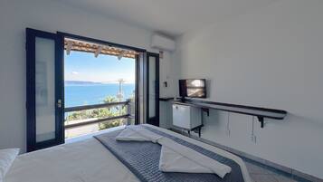 Un dormitorio con cama, televisor, un refrigerador pequeño y un balcón con vista al mar.