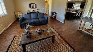 Amplia sala de estar con un sofá azul oscuro, una mesa de centro de madera con objetos decorativos y una zona de cocina al fondo.