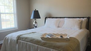 Una cama bien hecha con edredón blanco y almohadas, una lámpara de noche y una bandeja con un libro y un control remoto.