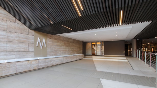 A spacious lobby with a modern design, featuring a prominent 'M' sign on the wall, a tiled floor, and a ceiling with linear lighting. - Sandton Smart Apartment Fifteen (Sandton)