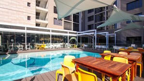 A poolside area with wooden tables and yellow chairs, surrounded by glass windows and a building with balconies. - Sandton Smart Apartment Fifteen (Sandton)