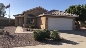 A residential house with a beige exterior, a white garage door, and a small front yard with gravel and a few shrubs. - Lavender Lane (Phoenix)