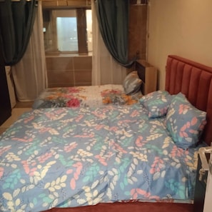 A bed with a floral-patterned bedspread, a red headboard, and a window with curtains. - Ali Jasem Group (Doha)