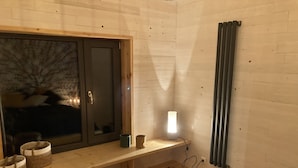 Een knus ingerichte kamer met een groot raam, een houten bankje, een nachtkastje met lamp en een radiator.