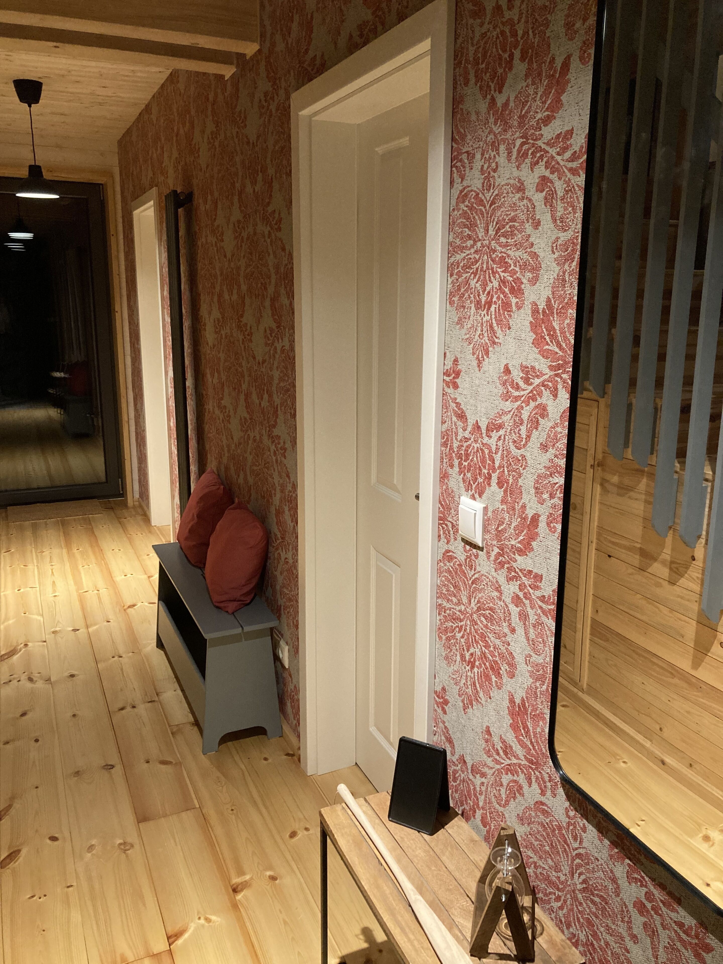 Un couloir doté d’un plancher en bois, d’un banc avec des coussins rouges, d’une porte et d’un mur orné d’un motif floral.