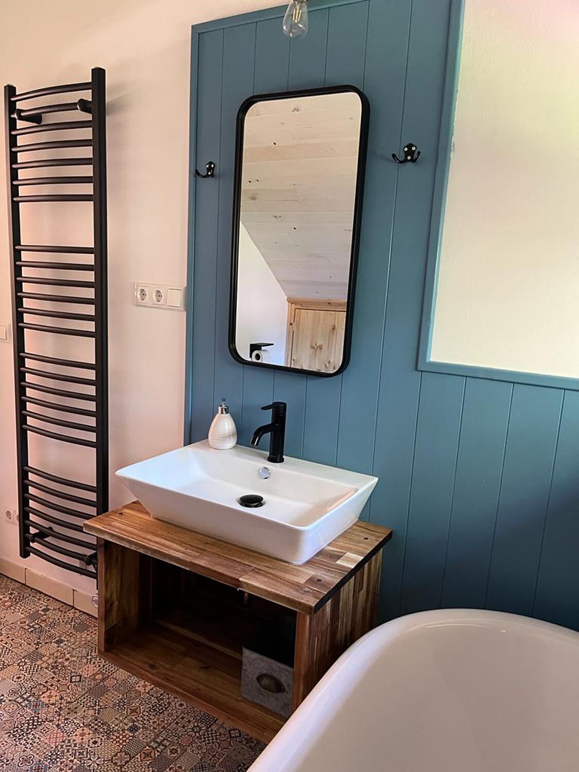 Een badkamer met een wit wastafel, een zwarte kraan en een spiegel erboven. Links een handdoekhouder en rechts een bad.