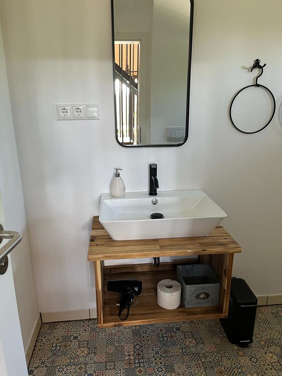 A bathroom with a white sink, a mirror, a towel ring, a toilet paper holder, and a trash bin.