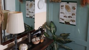 Une pièce avec une table d’appoint en bois sur laquelle se trouvent une lampe, un petit bol et des objets décoratifs. Un miroir est accroché au mur, au-dessus de la table. Le mur est orné d’une porte verte avec trois motifs circulaires et d’un panneau d’affichage où sont punaisées des feuilles de papier.