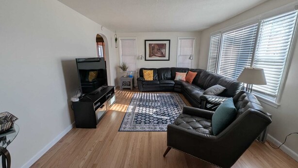 A living room with a black leather sofa, a black entertainment unit, a patterned rug, and a large window with blinds. - Trailview: Walk to Stadium! Free Fubo for World Cup, Plenty of Parking!  Dogs OK (Raytown)