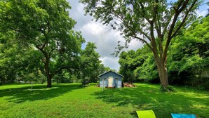 A backyard with a small blue shed, a green lawn, and a tree-lined area. - Trailview: Walk to Stadium! Free Fubo for World Cup, Plenty of Parking!  Dogs OK (Raytown)