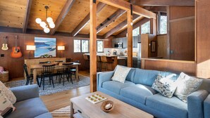 A spacious living area with a wooden ceiling, a blue sofa, a dining table, and a kitchen in the background.