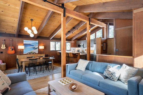A spacious living area with a wooden ceiling, a blue sofa, a dining table, and a kitchen in the background.