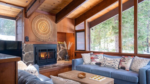 A cozy living room with a stone fireplace, a blue sofa with patterned cushions, and large windows offering a view of the forest.