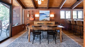 A wooden dining area with a large table and chairs, a painting on the wall, and a view through large windows.