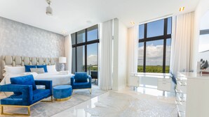 Un dormitorio moderno con una cama grande, sillones tapizados en azul, un aparador blanco y ventanas amplias con vistas al exterior.