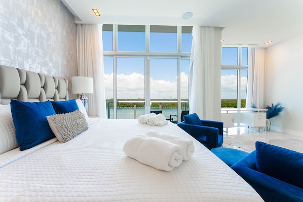 Una habitación de hotel moderna con una cama grande, almohadas azules y blancas, un escritorio y vistas al exterior.