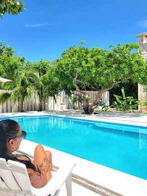 A swimming pool with a white deck, surrounded by lush greenery and a stone building.