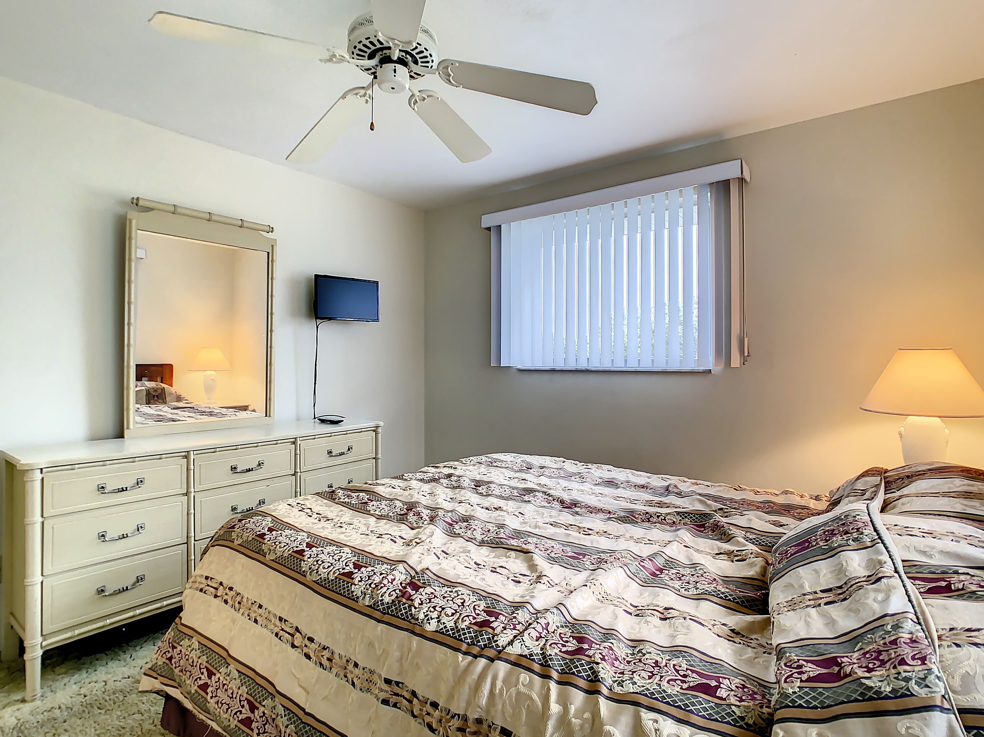 Une chambre à coucher comprenant un lit, une commode, un téléviseur, un ventilateur de plafond et une fenêtre avec des stores.