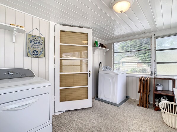 A laundry room with a front-loading washer and dryer, a wooden door, a window with blinds, and a shelf with laundry supplies.