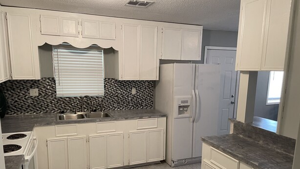 A kitchen with white cabinets, a granite countertop, a built-in stove, a refrigerator, and a backsplash with a black and white tile pattern. - Mark and Bobby’s Island (Wharton)