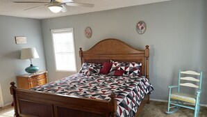 A bedroom with a wooden bed, a nightstand with a lamp, a chair, a window with blinds, and a ceiling fan. - Mark and Bobby’s Island (Wharton)